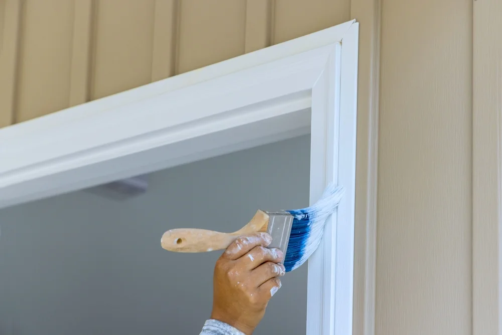 Painter applying white paint to door frame trim with paintbrush