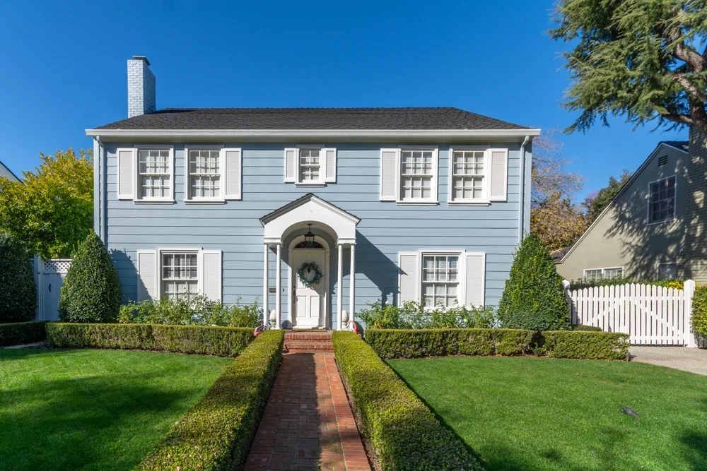 Two-story light blue colonial house with white shutters and red brick walkway