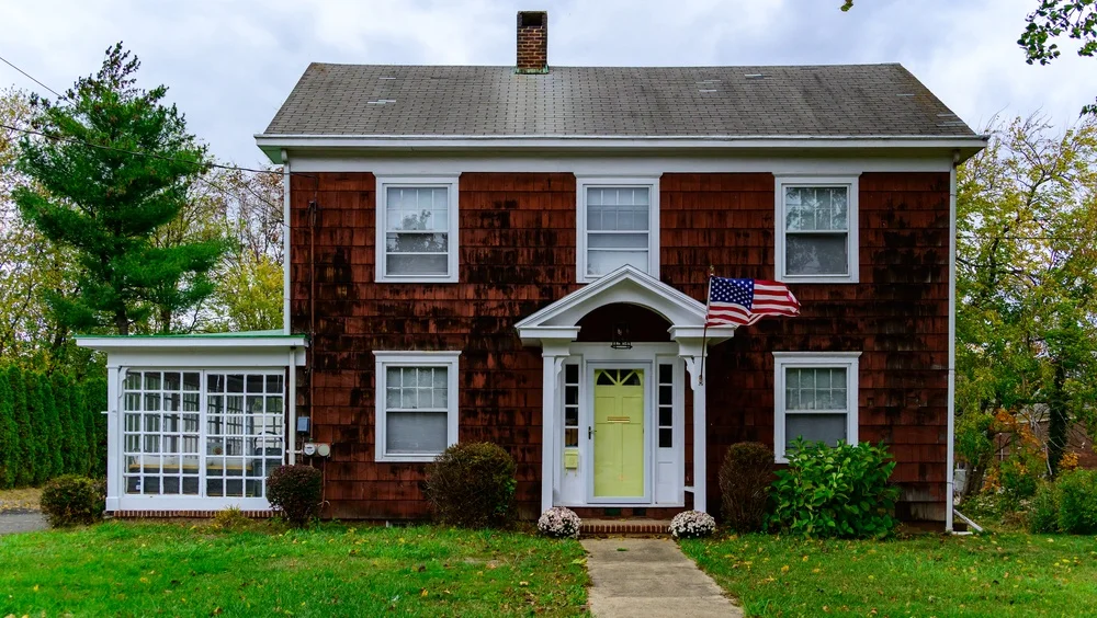Two-story brown shingle house with a pale yellow front door, white trim, and an American flag attached to the porch.
